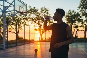 negro hombre haciendo Deportes en mañana, Bebiendo agua en baloncesto Corte en amanecer foto