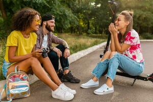 happy young company of smiling friends sitting in park on grass with electric kick scooter photo