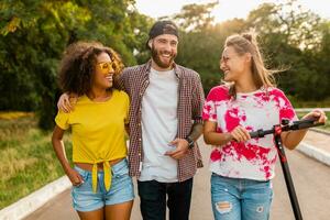 happy young company of smiling friends walking in park with electric kick scooter photo