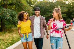 happy young company of smiling friends walking in park with electric kick scooter photo