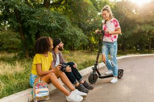 happy young company of smiling friends sitting in park on grass with electric kick scooter photo