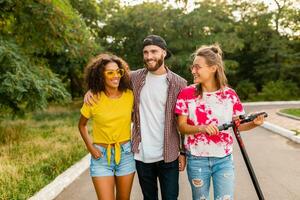 happy young company of smiling friends walking in park with electric kick scooter photo