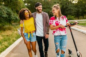 happy young company of smiling friends walking in park with electric kick scooter photo