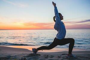 young woman doing sport exercises on sunrise beach in morning photo