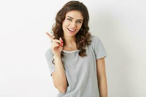 close up portrait of attractive young woman smiling face expression holding finger up, showing aside, looking in camera, natural beauty, t-shirt, white teeth, isolated on white background, gesturing photo