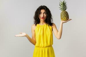 retrato de joven sorprendido bonito mujer en amarillo vestido, participación piña, gracioso emoción, conmocionado cara expresión, verano estilo, Fruta dieta, mirando en cámara, mezclado carrera, aislado, participación manos arriba foto