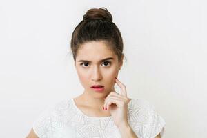 close-up portrait of young thinking woman in white blouse biting her lip, frowning, holding finger at her face, emotional, serious face expression, looking into camera, having a problem isolated photo