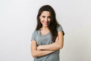 retrato de elegante joven bonito mujer sonriente en gris camiseta en blanco estudio fondo, aislado, natural mirar, largo marrón cabello, cruzado brazos foto