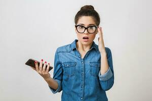 shocked young woman holding smart phone, wearing stylish glasses, opened mouth, looking into camera, surprised face expression, isolated on white background, denim shirt photo