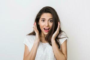 portrait of young surprised woman in white blouse with long hair, positive shocked face expression, happy, holding hands up at her face, isolated on studio background photo