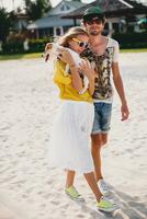 hipster young stylish hipster couple in love walking playing dog puppy jack russell in tropical beach, white sand, cool outfit, romantic mood, having fun, sunny, man woman together, vacation photo