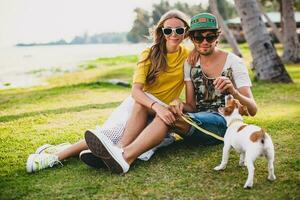 young stylish hipster couple in love sitting on grass playing dog puppy jack russell in tropical beach, white sand, cool outfit, romantic mood, having fun, sunny, man woman together, vacation photo