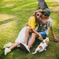 young stylish hipster couple in love sitting on grass playing dog puppy jack russell in tropical beach, white sand, cool outfit, romantic mood, having fun, sunny, man woman together, vacation photo