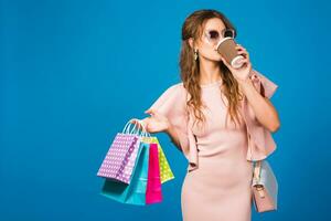 pretty young stylish sexy woman in pink luxury dress, summer fashion trend, chic style, sunglasses, blue studio background, shopping, holding paper bags, drinking coffee, shopaholic photo