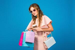 pretty young stylish sexy woman in pink luxury dress, summer fashion trend, chic style, sunglasses, blue studio background, shopping, holding paper bags, drinking coffee, shopaholic photo