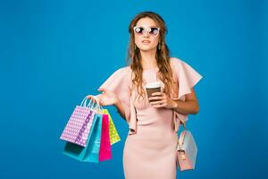 pretty young stylish sexy woman in pink luxury dress, summer fashion trend, chic style, sunglasses, blue studio background, shopping, holding paper bags, drinking coffee, shopaholic photo