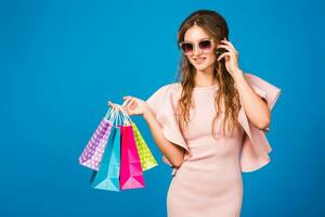 pretty young stylish sexy woman in pink luxury dress, summer fashion trend, chic style, sunglasses, blue studio background, shopping, holding paper bags, drinking coffee, shopaholic photo