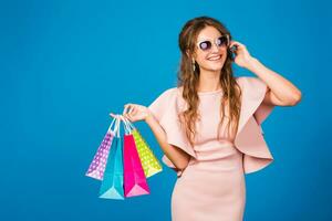 pretty young stylish sexy woman in pink luxury dress, summer fashion trend, chic style, sunglasses, blue studio background, shopping, holding paper bags, drinking coffee, shopaholic photo