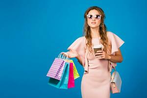 pretty young stylish sexy woman in pink luxury dress, summer fashion trend, chic style, sunglasses, blue studio background, shopping, holding paper bags, drinking coffee, shopaholic photo