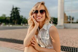 young attractive cute stylish blonde woman sitting in city street in summer fashion style photo