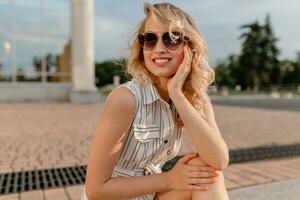 young attractive cute stylish blonde woman sitting in city street in summer fashion style photo