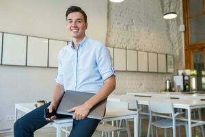 happy young attractive smiling man sitting in co-working open office, holding laptop photo