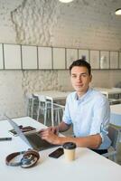 joven hermoso sonriente hombre sentado en abierto espacio oficina trabajando en ordenador portátil foto