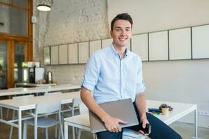 young attractive smiling man sitting in co-working open office, holding laptop photo