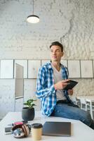 young handsome man in chekered shirt sitting on table using tablet computer in co-working office photo