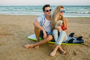 young smiling couple having fun on beach sitting on sand with surf boards playing with dog photo