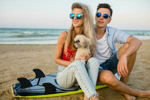 young smiling couple having fun on beach sitting on sand with surf boards playing with dog photo