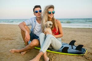 young smiling couple having fun on beach sitting on sand with surf boards playing with dog photo