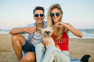 young smiling couple having fun on beach sitting on sand with surf boards playing with dog photo