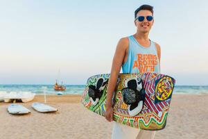 young athletic man with kite surfing board posing on beach wearing sunglasses on summer vacation, active sports hobby photo