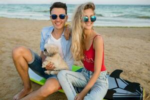 young smiling couple having fun on beach sitting on sand with surf boards playing with dog photo