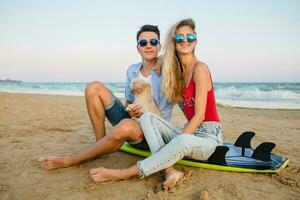 young smiling couple having fun on beach sitting on sand with surf boards playing with dog photo