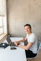 joven hermoso hombre en casual atuendo sentado a mesa trabajando en computadora portátil, mecanografía, persona de libre dedicación a hogar foto