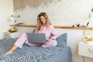 young pretty blond woman in pink pajamas sitting on bed working on laptop, freelancer at home photo