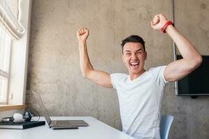 young handsome smiling man in casual outfit sitting at table working on laptop photo