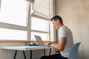 joven hermoso sonriente hombre en casual atuendo sentado a mesa trabajando en ordenador portátil foto