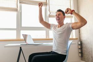 young handsome smiling man in casual outfit sitting at table working on laptop photo