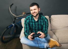 young hipster handsome bearded man sitting on couch at home, playing video game photo