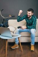 young hipster handsome bearded man sitting on couch at home, playing video game photo