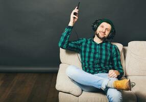 joven hipster hermoso barbado hombre sentado en un sofá a hogar, escuchando a música en auriculares foto