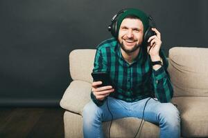 young hipster handsome bearded man sitting on a couch at home, listening to music on headphones photo