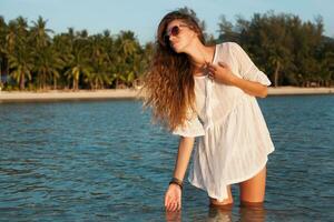 slim beautiful woman in white cotton dress walking on tropical beach photo