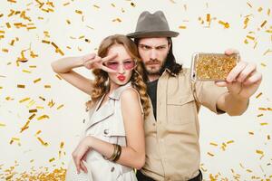 young stylish hipster couple in love making self photo, celebrating disco party, having fun, amazed, exited, funny face, golden confetti, holding phone, trendy apparel, cruise style, white background photo