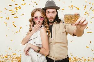 young stylish hipster couple in love making self photo, celebrating disco party, having fun, amazed, exited, funny face, golden confetti, holding phone, trendy apparel, cruise style, white background photo