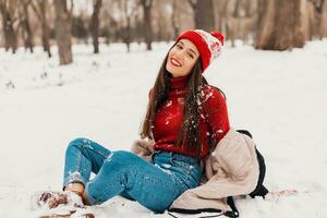 young pretty smiling happy woman in red mittens and knitted hat wearing winter coat sitting on snow in park, warm clothes photo