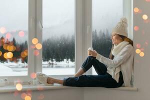 young attractive woman in stylish white knitted sweater, scarf and hat sitting at home on windowsill at Christmas holding glass snow ball present decoration, winter forest view, lights bokeh photo